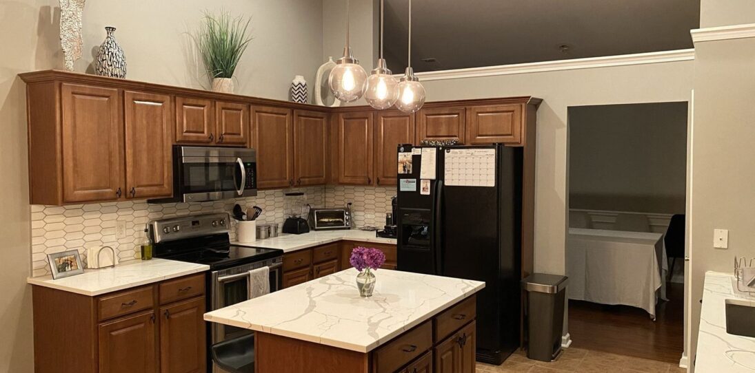 Wide view of a kitchen with honey oak cabinets, calacatta quartz counters, three glass pendant lights, and a vaulted ceiling before the paint renovation began.