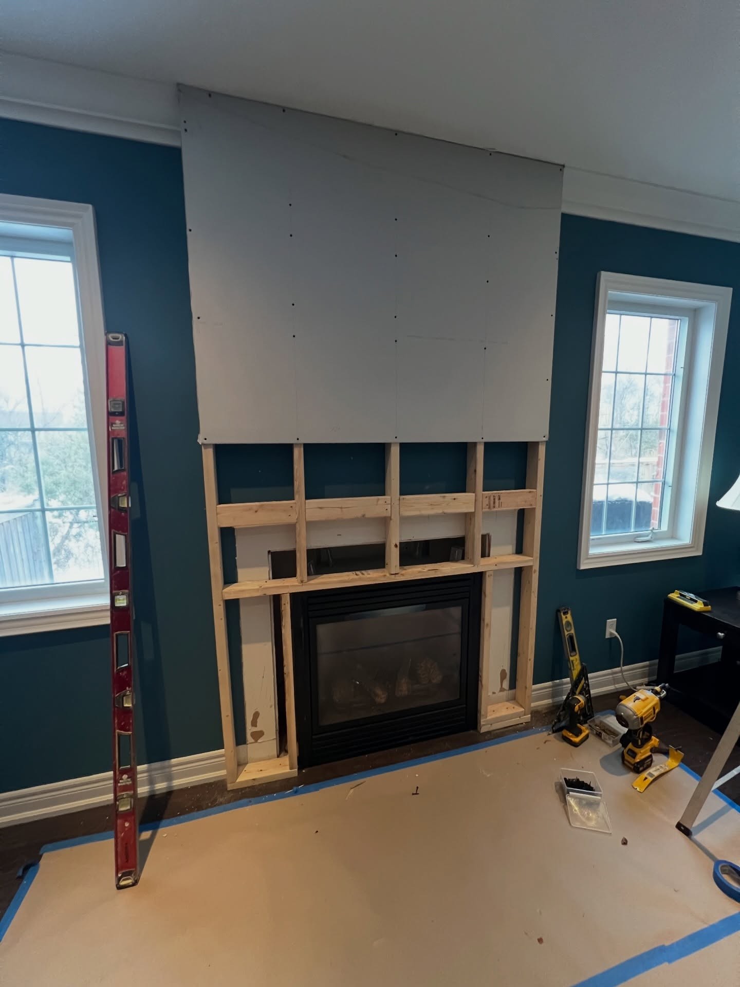 Fireplace wall with most of the drywall installed, exposed framing around the firebox opening, and tools visible on the floor including a level and ladder.