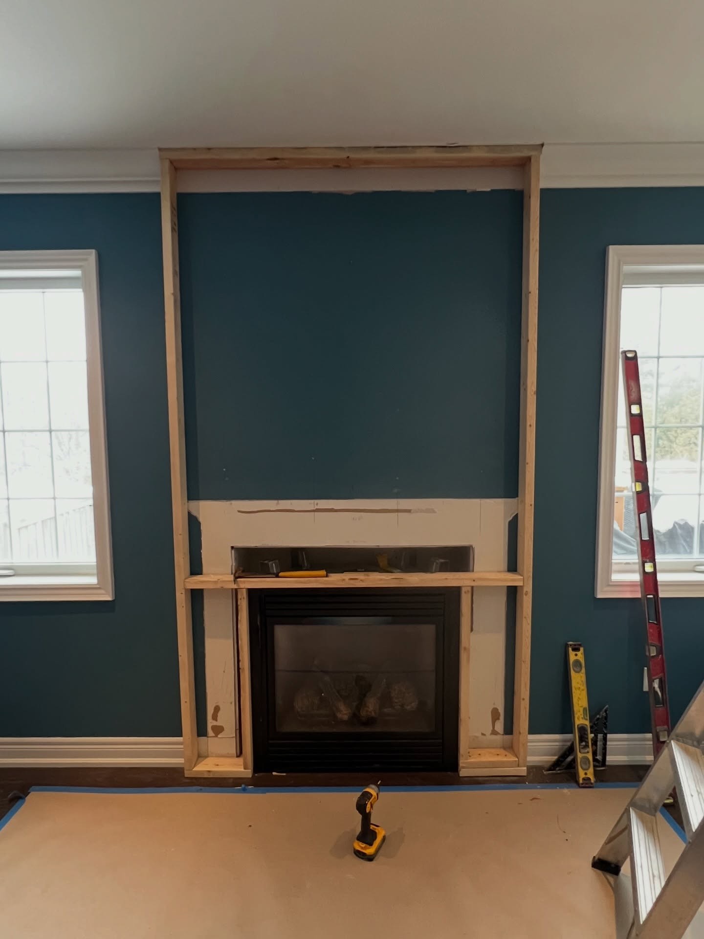 Completed vertical framing between the two windows showing the new fireplace wall will reach all the way to the ceiling, with a drill on the floor and construction paper taped down.