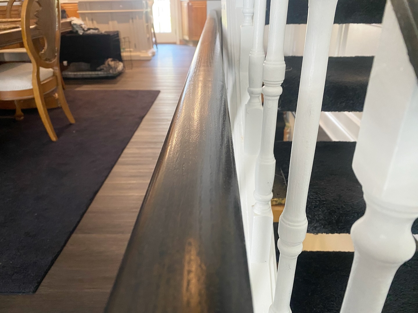 Side profile of the dark stained handrail with white spindles, dark wood dining room floor and a dark area rug in the background.