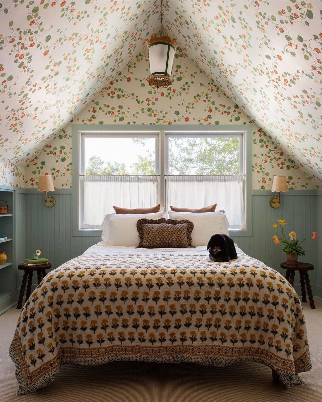 Attic bedroom with orange and green floral wallpaper covering the sloped ceiling, sage green beadboard wainscoting, yellow block print quilt, and a black puppy on the bed