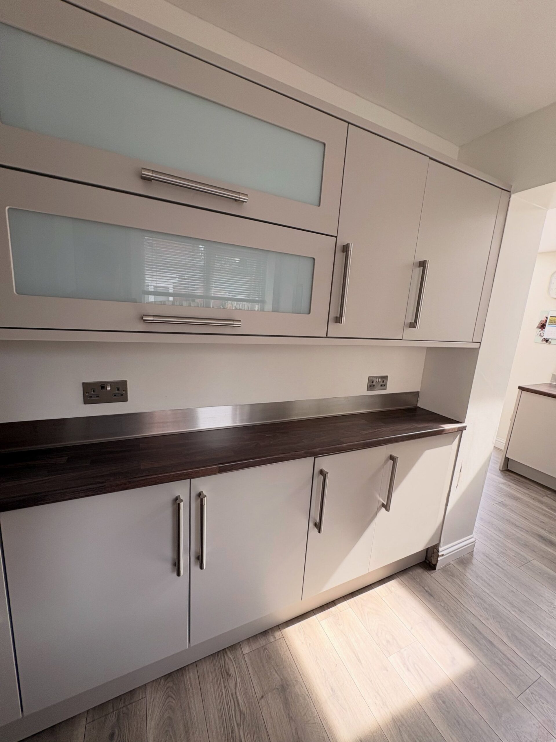 Wide view of upper and lower kitchen cabinets freshly sprayed in Purbeck Stone, two frosted glass doors in the center with new shorter brushed nickel handles, dark walnut counter below.