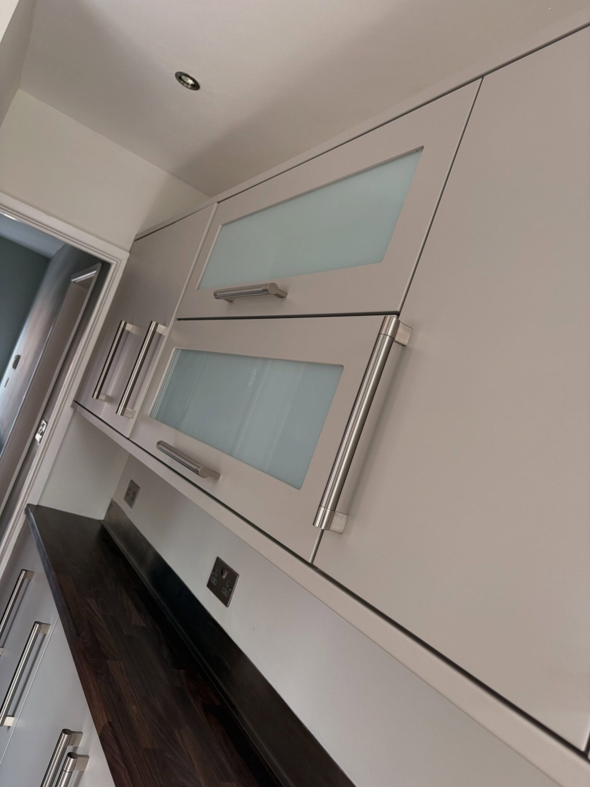 Close up of two frosted glass cabinet doors sprayed in Purbeck Stone with shorter brushed nickel handles, dark walnut counter and matching lower cabinet handles visible below.