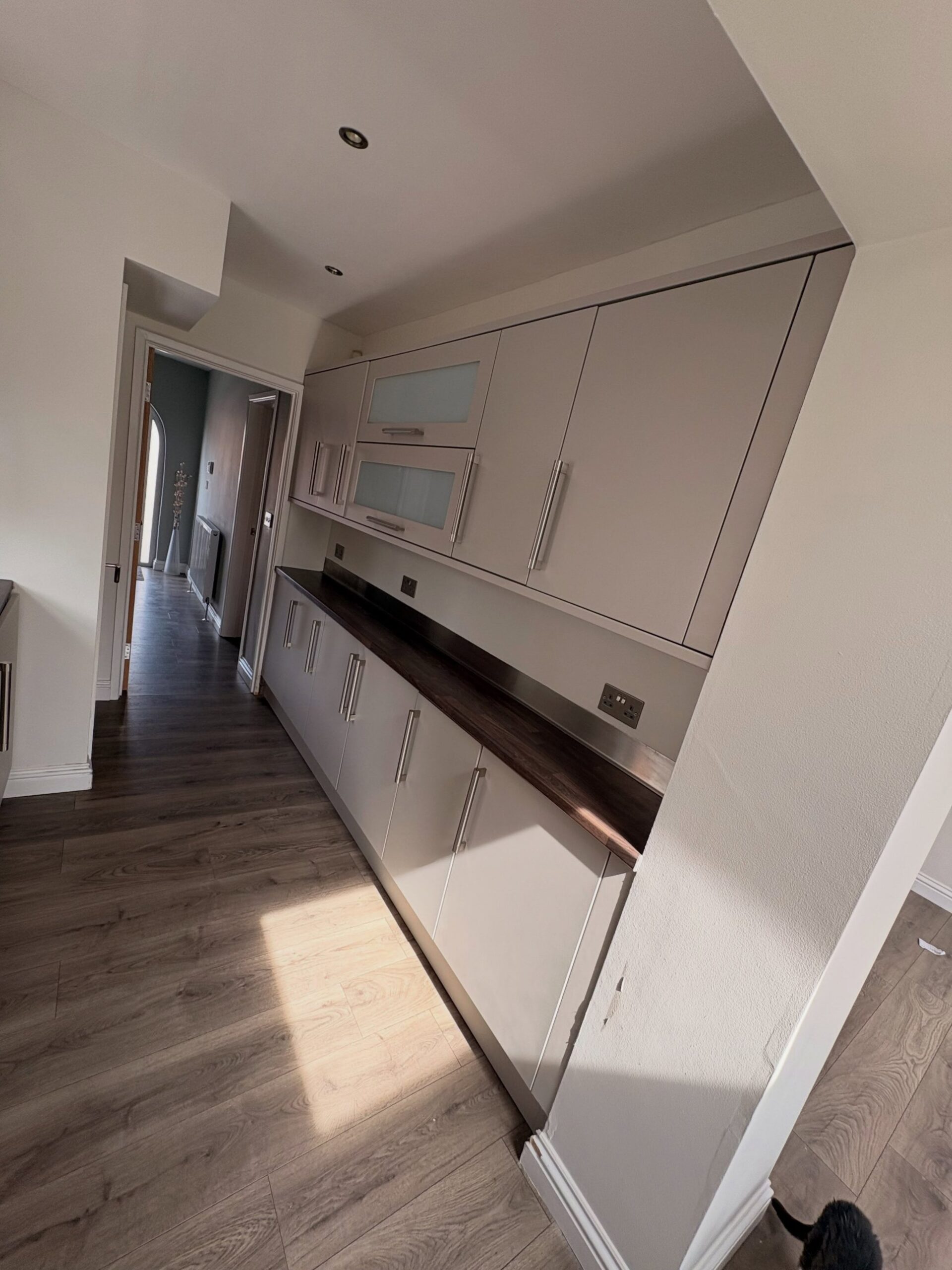 Finished galley viewed from the hallway, cabinets sprayed in Purbeck Stone with frosted glass detail, dark walnut counter and shorter brushed nickel handles, hallway and vase visible in the background.