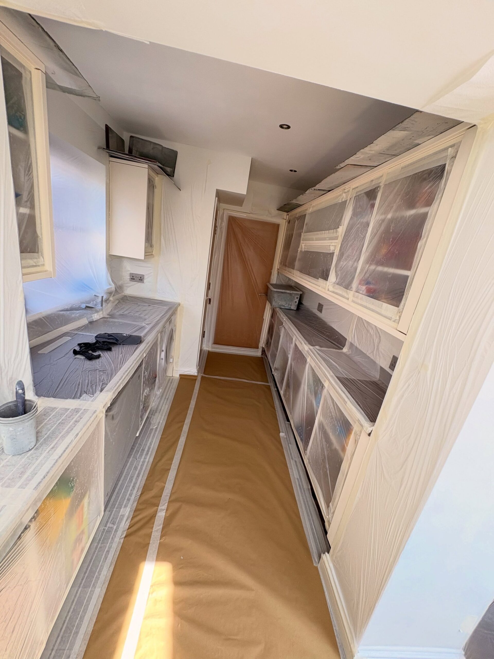 Narrow galley kitchen fully covered in clear plastic sheeting and masking paper on the floor, cabinets and counters wrapped for a spray job, afternoon light coming through the window.