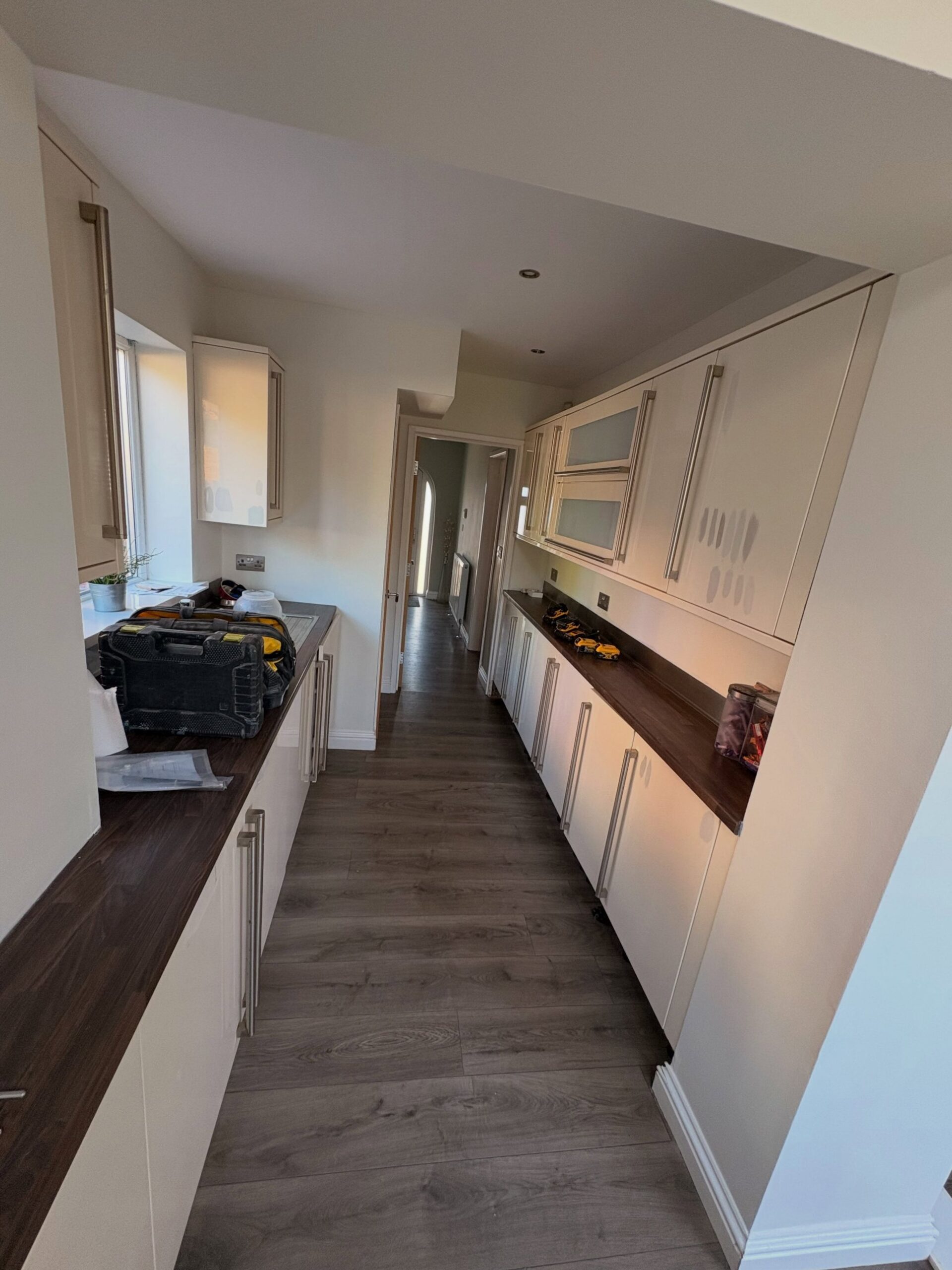 Galley kitchen viewed down the hallway, cream cabinets with grey patch marks on the upper doors, dark walnut counters, long vertical brushed bar handles throughout.