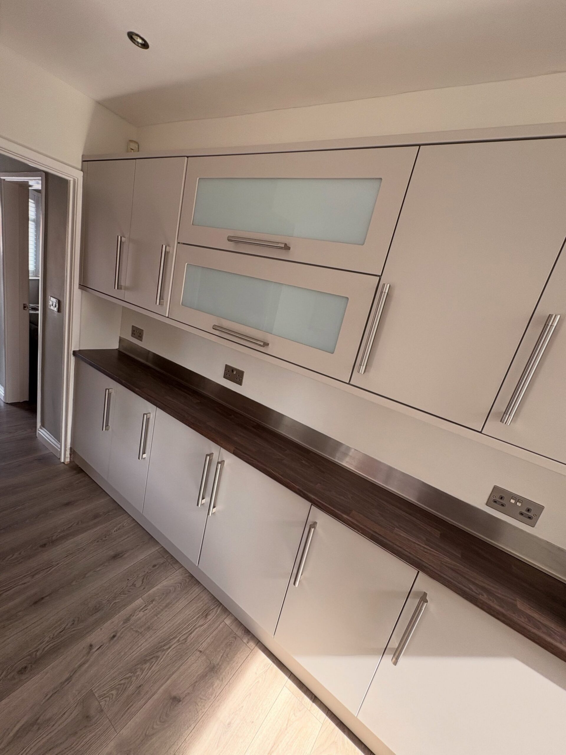 Finished galley kitchen wall sprayed in Farrow and Ball Purbeck Stone, upper cabinets with frosted glass panels and shorter brushed nickel handles, dark walnut counter running the length of the wall.