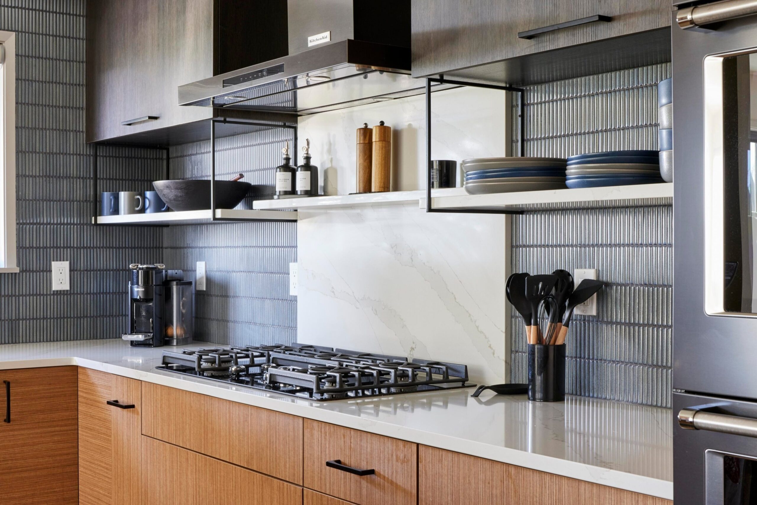 Close view of a gas cooktop with cast iron grates set into a white quartz counter, with vertical blue kit-kat tile on the wall, a stainless hood above, and open metal shelves holding mugs, oils, and a wood bowl.