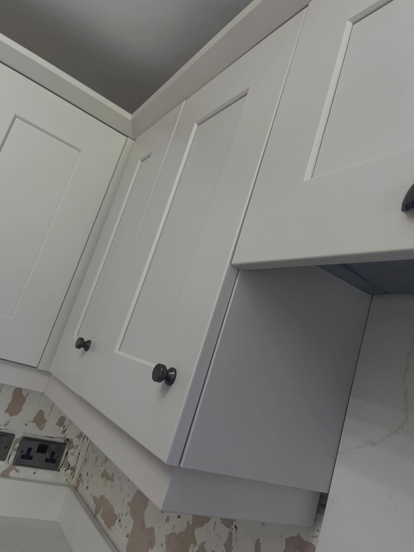 Close up of two shaker cabinet doors painted in matte Gentle Fawn with small round burnt steel knobs, set against a lightly scuffed plaster wall.