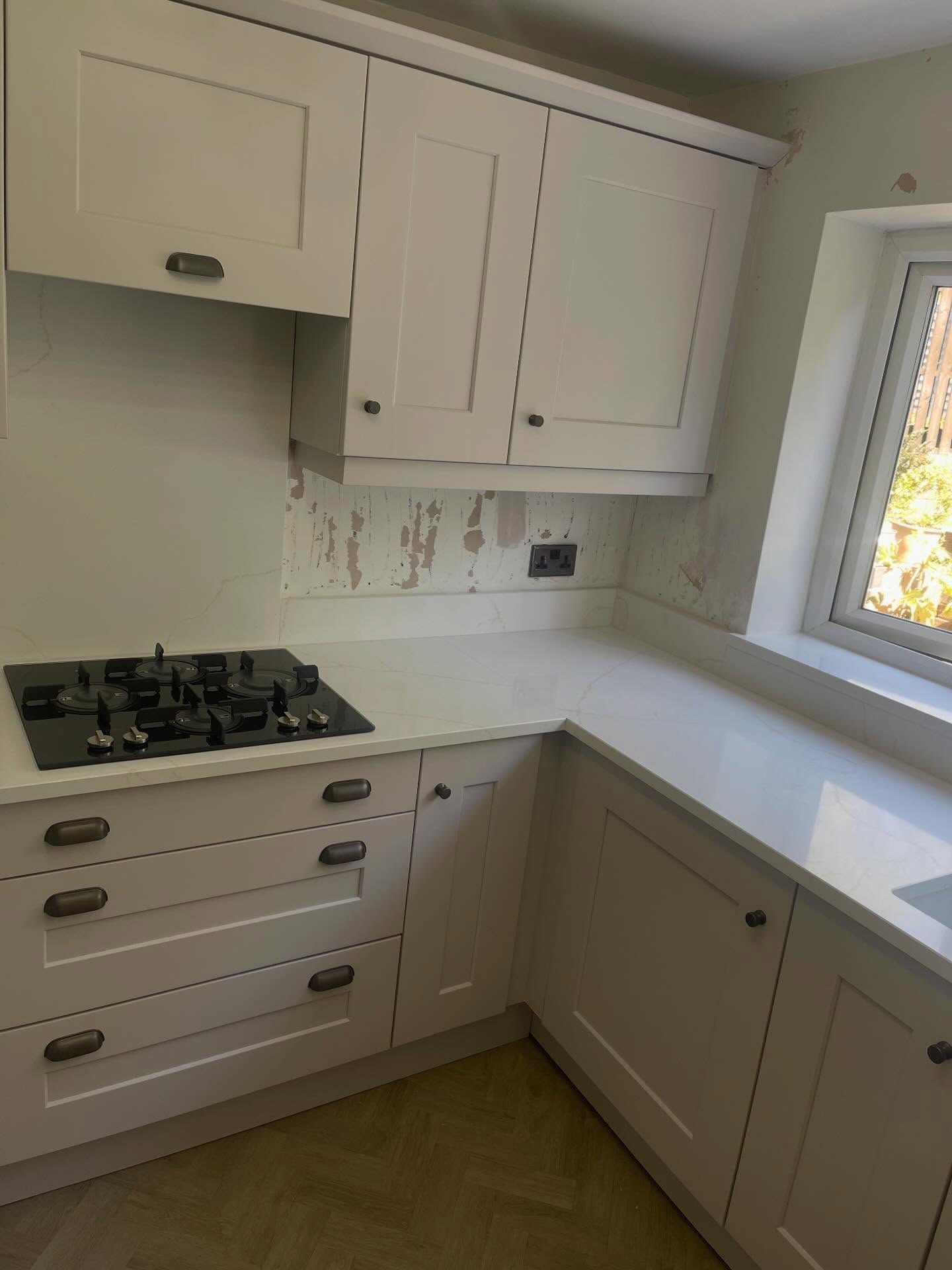 Painted Gentle Fawn kitchen with a black gas hob set into creamy veined quartz counter, soft matte cabinet doors, burnt steel cup handles on the drawers and a window to the garden.