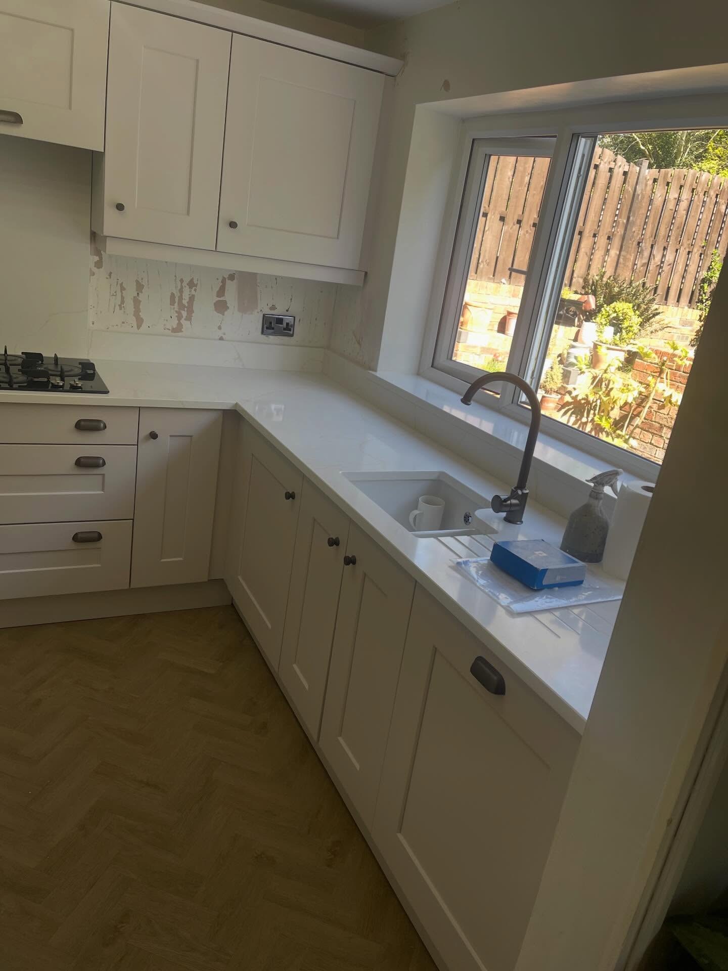 Painted sink run in soft Gentle Fawn with a white undermount sink, matte black gooseneck tap, creamy quartz worktop with carved drainer grooves and herringbone wood-look floor.