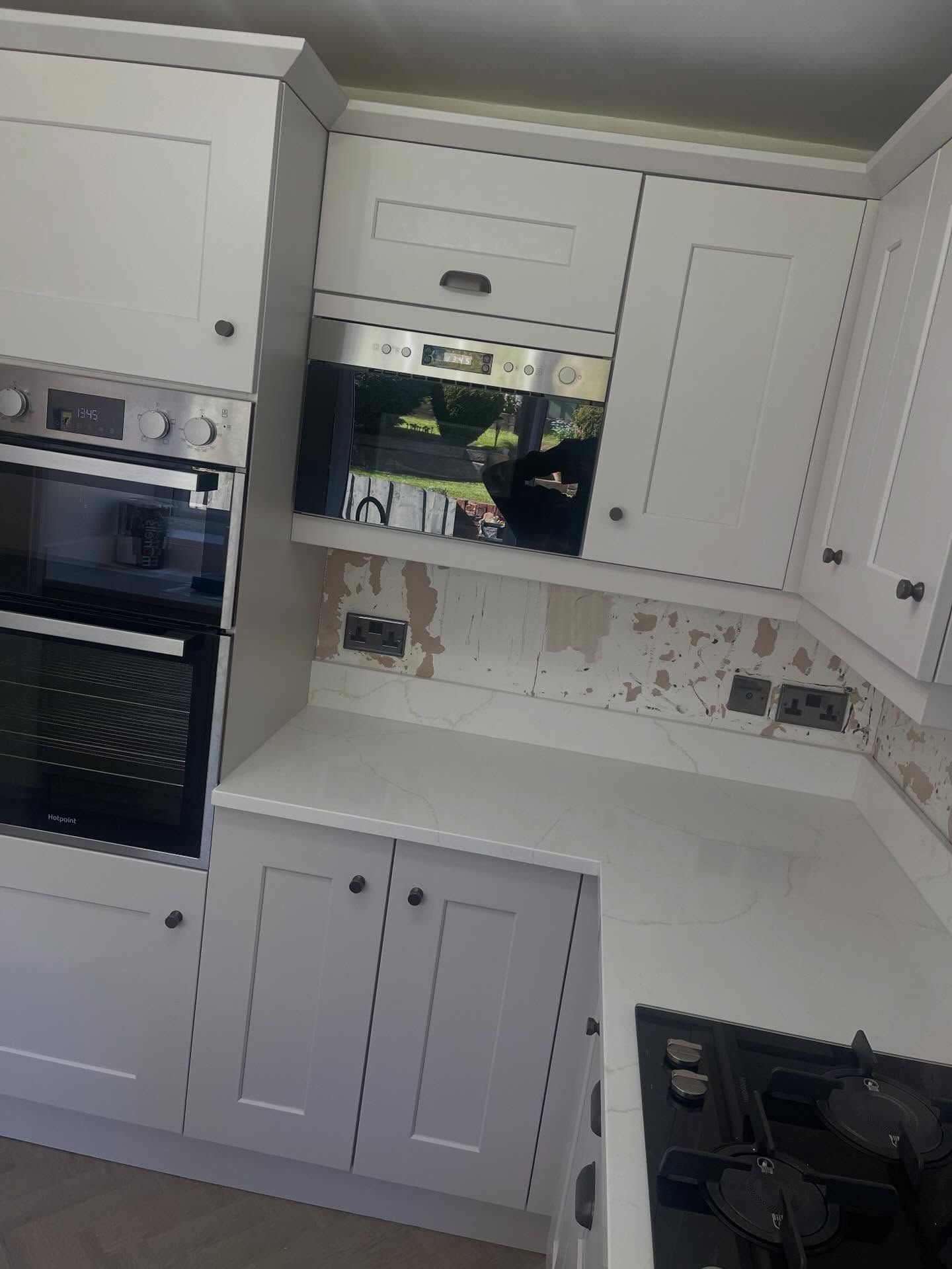 Finished kitchen corner showing a built in microwave with mirrored door reflecting the garden, Gentle Fawn upper and lower cabinets, burnt steel knobs and a quartz counter with original plaster patches still showing on the backsplash wall.