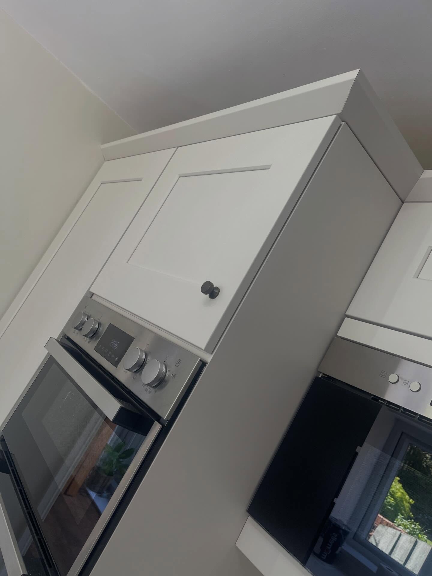 Angled corner view of the Gentle Fawn kitchen showing the oven stack beside a tall end panel, soft matte doors, and burnt steel knobs reading quietly against the paint.