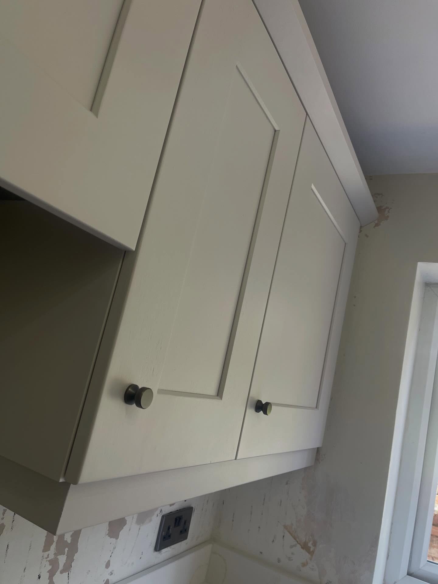 Close up angled view of Gentle Fawn upper cabinets wrapping into a corner, with the warm matte paint catching daylight and small burnt steel knobs on each door.