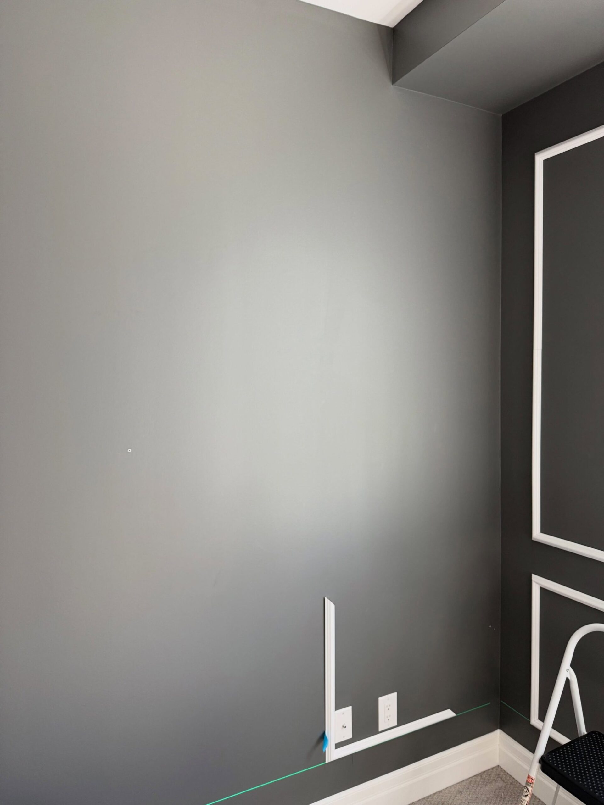 A gray office wall mostly empty, with a loose piece of white baseboard pulled off at the bottom and a green laser line projected across the lower wall at baseboard height during paint prep.