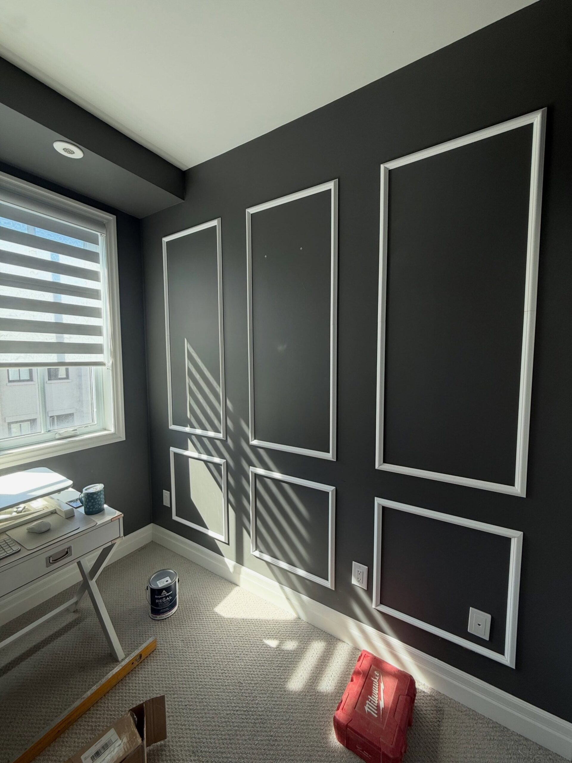 A small home office with dark charcoal walls, bright white panel molding on the back wall, a white campaign desk by the window, a Milwaukee tool box and a Benjamin Moore paint can on the carpet.