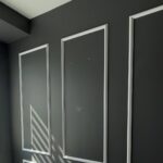 Dark charcoal home office wall with three tall white panel moldings on the back wall, a window with zebra blinds and white casing on the left, a white ceiling edge above, and sunlight casting diagonal shadow stripes across one panel.