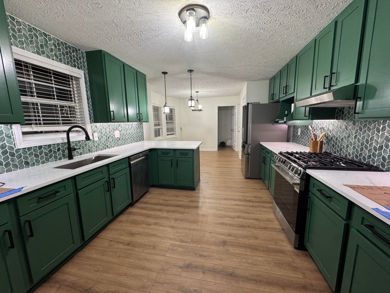 Wide daylight view of the finished green kitchen remodel with Sherwin-Williams Kale cabinets, green leaf tile backsplash wrapped around the window frame, white quartz countertops, Sapelo Shore LVP flooring, a stainless gas range, and black metal pendant lights.