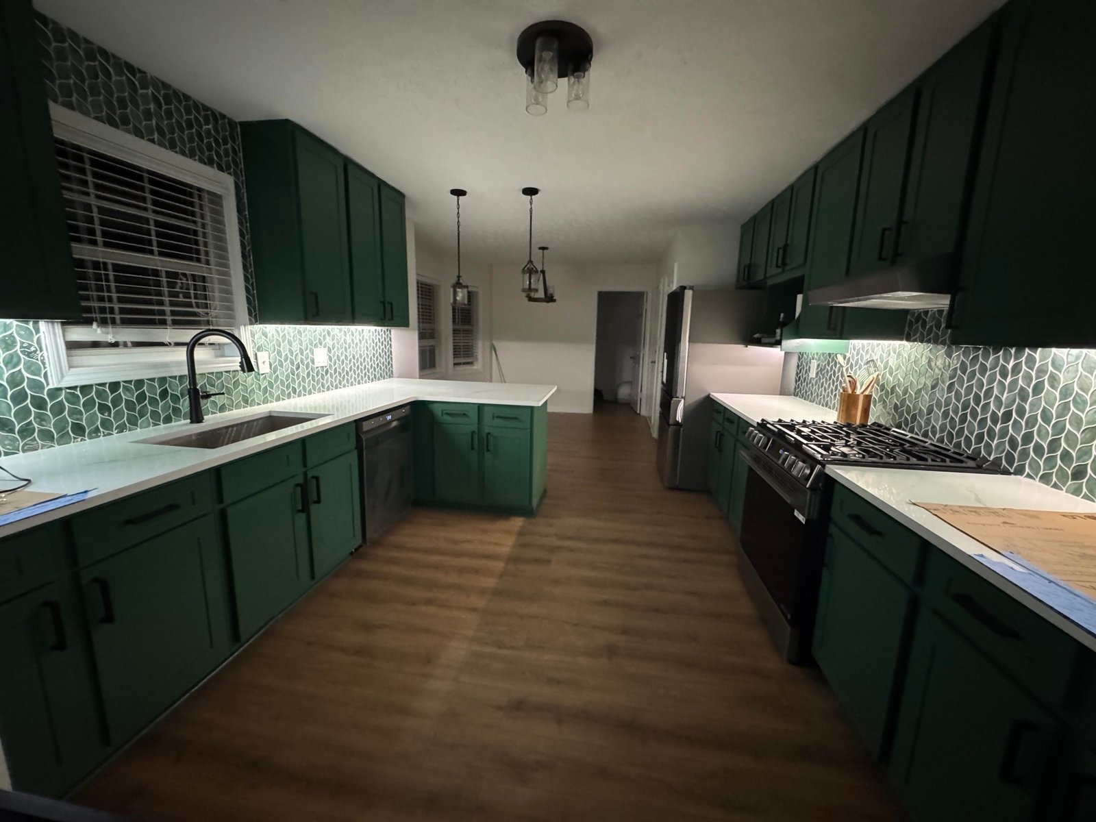 After view of the finished kitchen showing deep green Sherwin-Williams Kale painted cabinets, a green leaf-shaped mosaic tile backsplash with white grout, white quartz countertops, a matte black pull-down faucet, black hardware, and LVP wood-look flooring under warm pendant lighting.