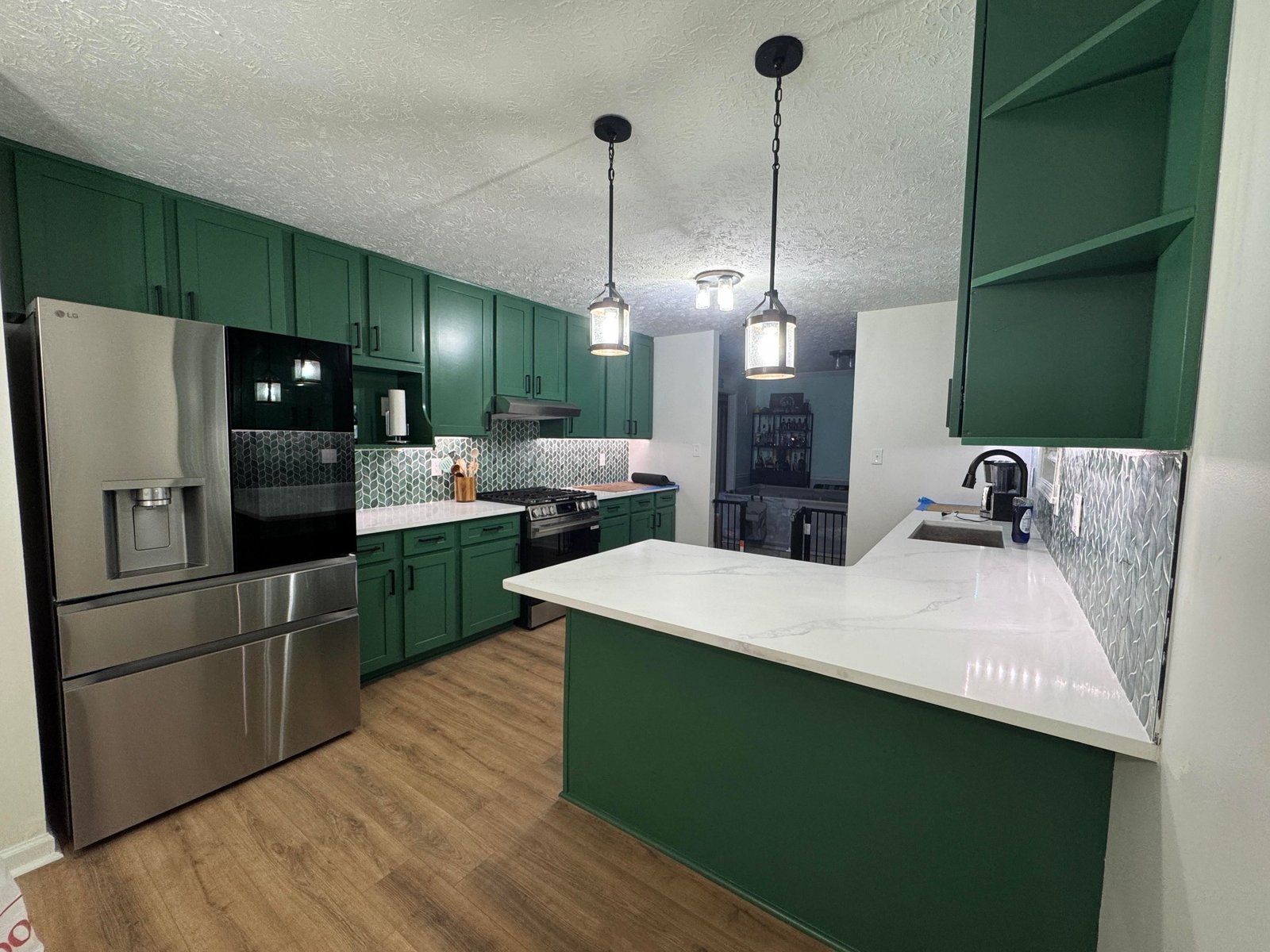 Kitchen peninsula and bar view showing green Kale cabinets, an open shelf cutout at the end, a white quartz waterfall countertop, a stainless LG French-door fridge, black pendant lights, and LVP wood flooring leading into the dining area.