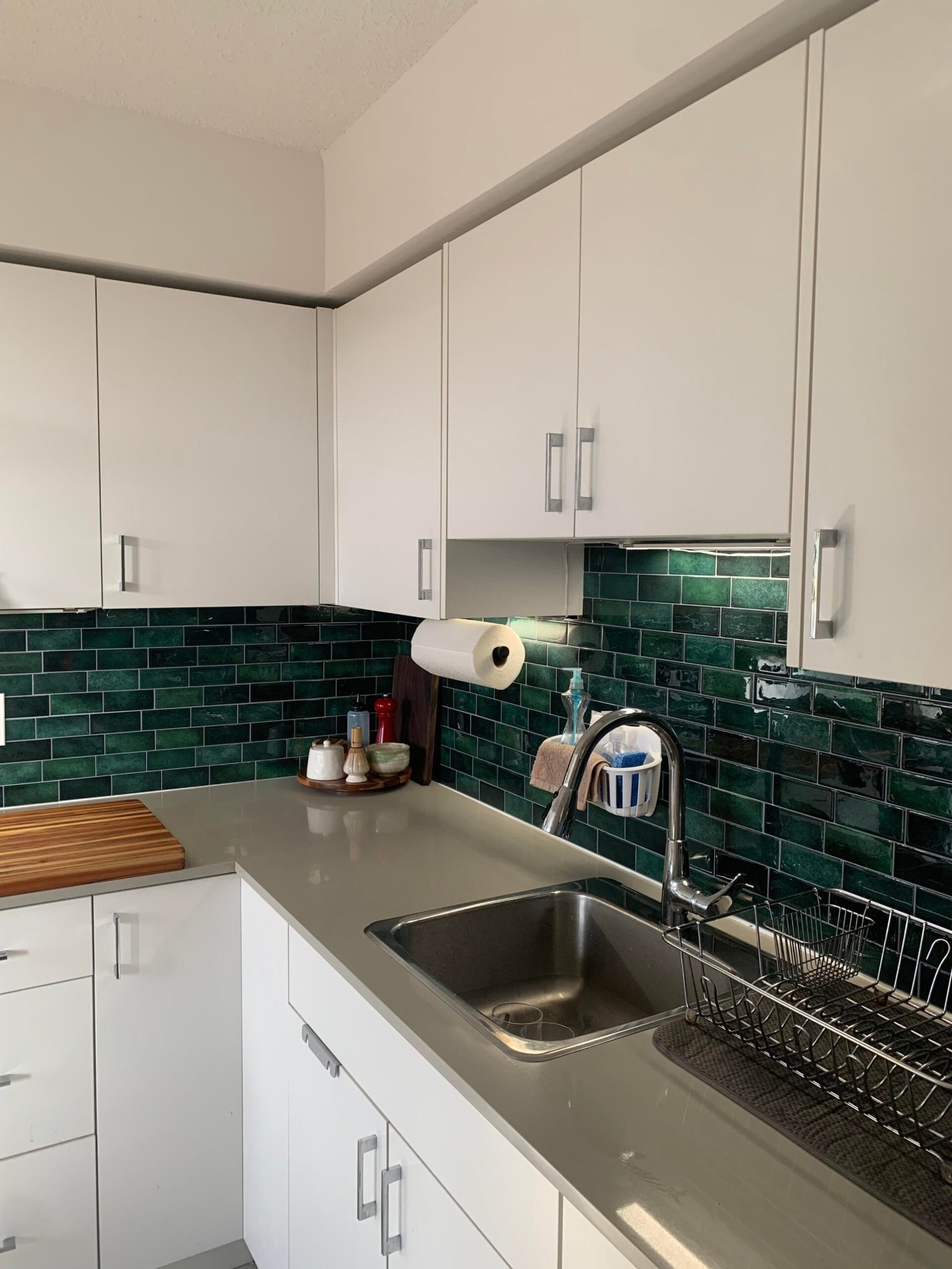 Finished rental kitchen corner with a glossy dark green subway tile peel-and-stick backsplash wrapping around the sink wall, white cabinets above, gray quartz counter, and a thick teak butcher block cutting board on the left.