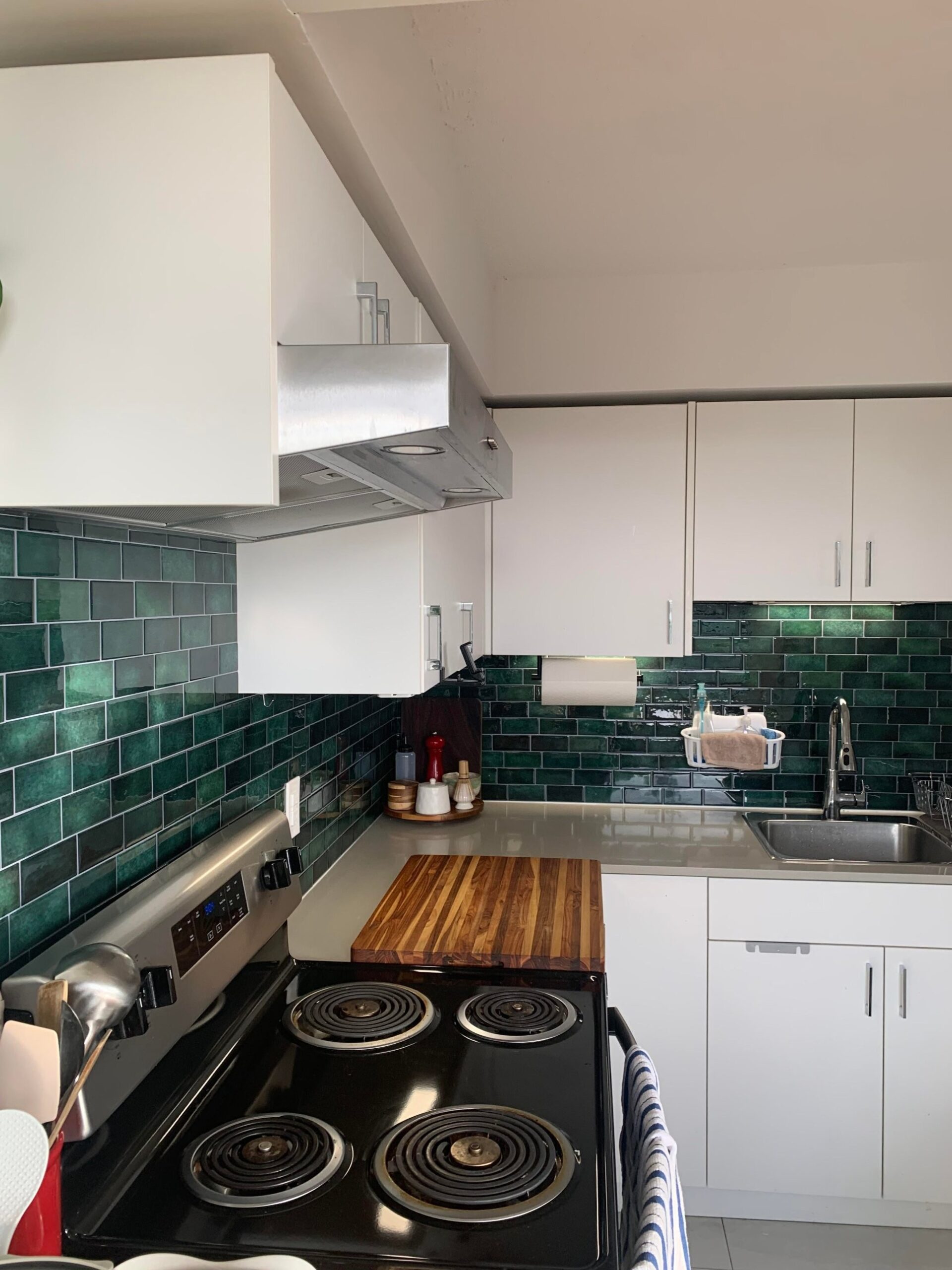 Rental kitchen with a black electric coil stove, stainless range hood, white upper cabinets, and a finished dark green peel-and-stick subway tile backsplash running from counter to upper cabinets around the stove and outlet.