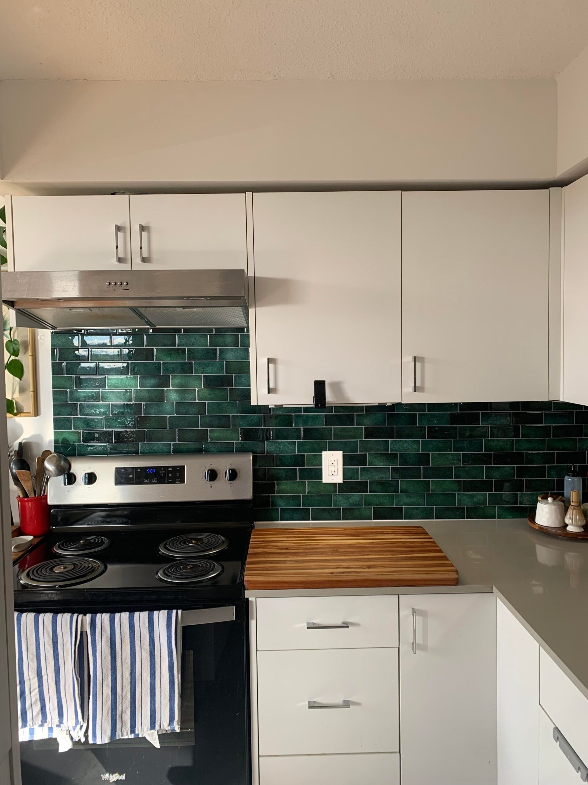 Rental kitchen stove area shot straight on with a thick teak butcher block on the gray quartz counter, finished dark green peel-and-stick subway tiles across the full backsplash, white cabinets above, and a plant on the far left.