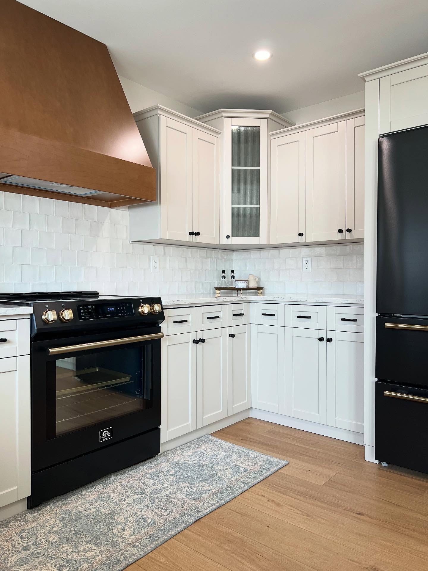 Corner view of the finished kitchen showing the warm wood hood on the left, a glass-front corner cabinet, soft greige Shaker cabinetry, white textured square backsplash tile, a matte black slide-in range with brushed brass knobs, a matte black bottom-freezer fridge with brass handle, and natural oak flooring with a patterned runner.