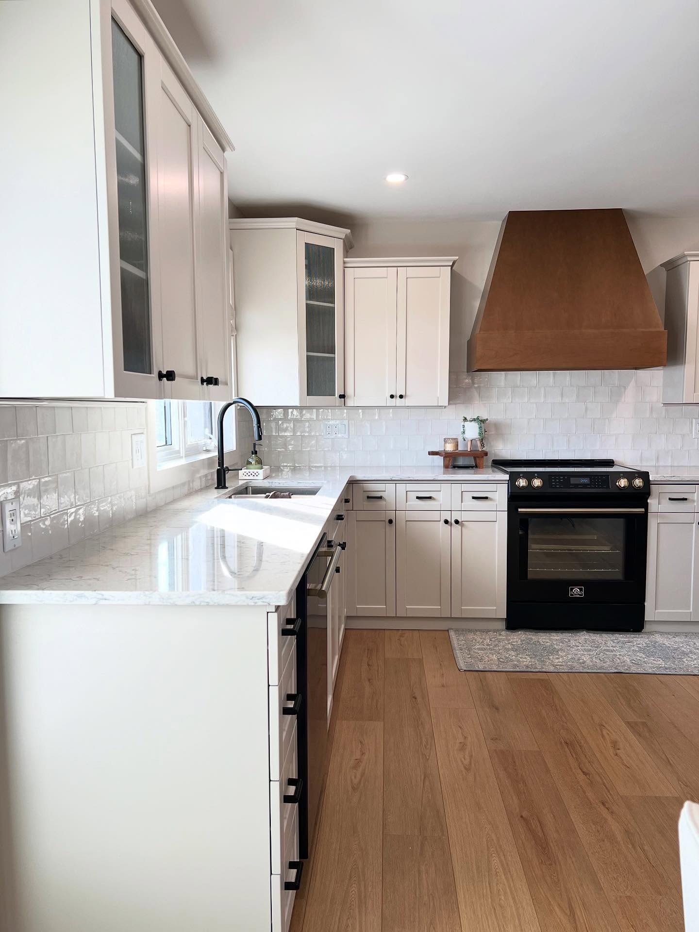 Wide view of a renovated U-shaped kitchen with soft greige Shaker cabinets, white quartz counters, white handmade-look square tile backsplash, a warm wood vent hood, a matte black range with brass accents, black cabinet hardware, and natural oak flooring.