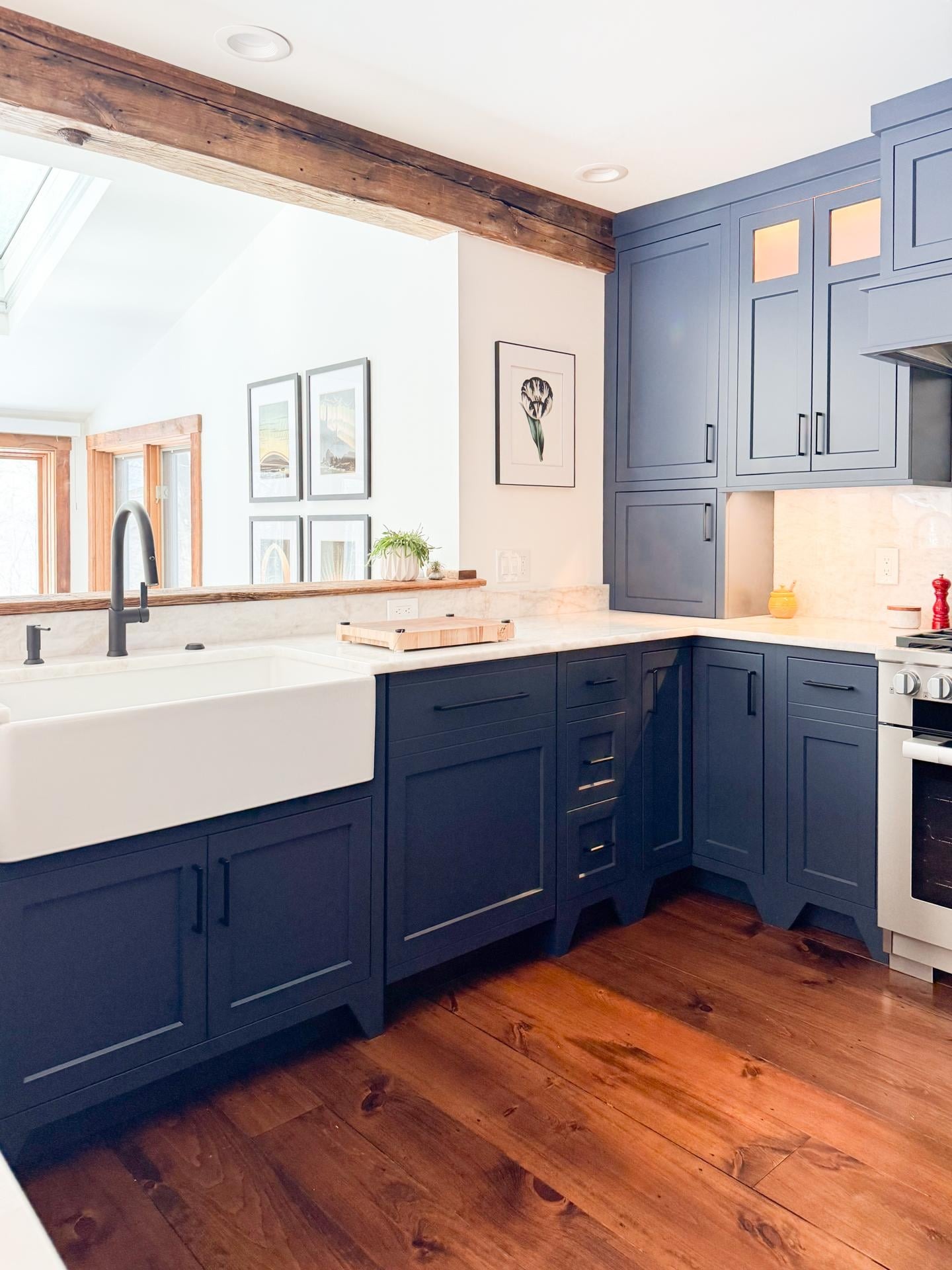 Corner view of the new kitchen with a white apron farmhouse sink, matte black pull-down faucet, framed botanical art, and a skylight bringing light down over navy cabinets.