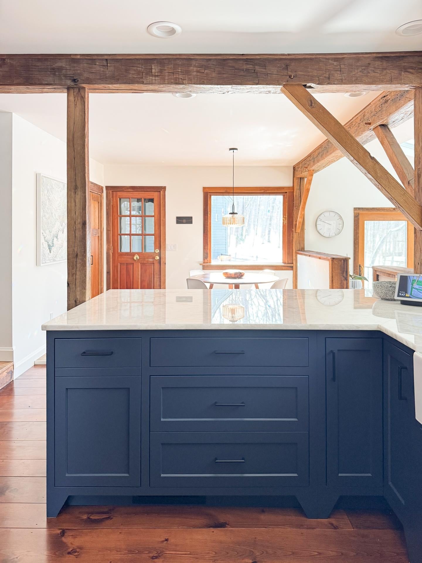 Navy peninsula with marble counter, matte black hardware, and a view through the beams to the dining nook and back door.
