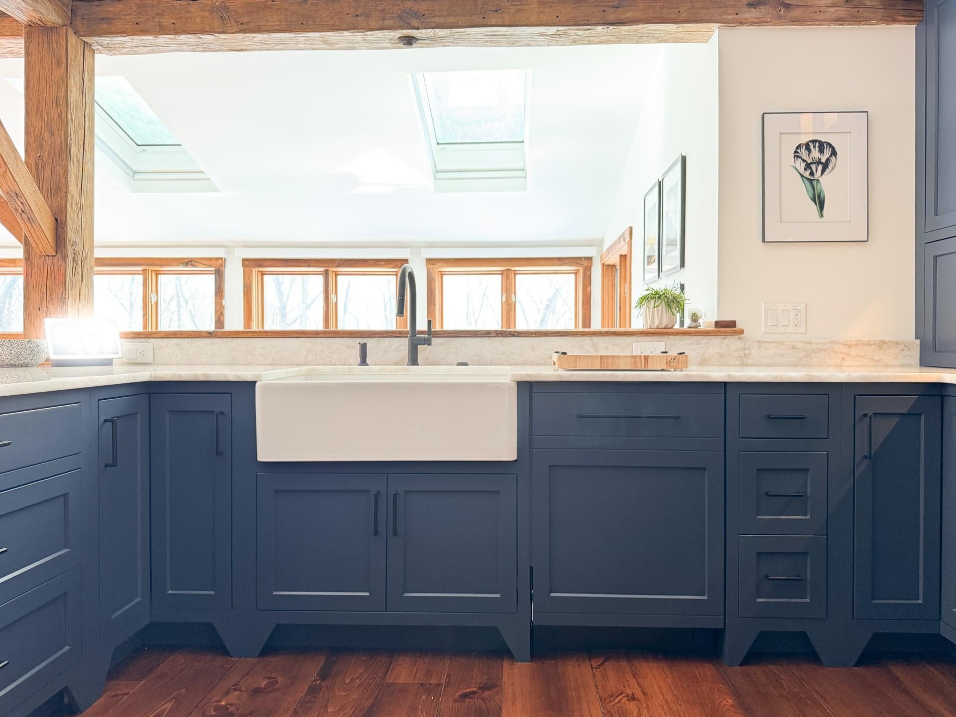 Farmhouse sink under a run of trim windows and twin skylights, with navy lower cabinets, marble counters, and heavy wood beams overhead.