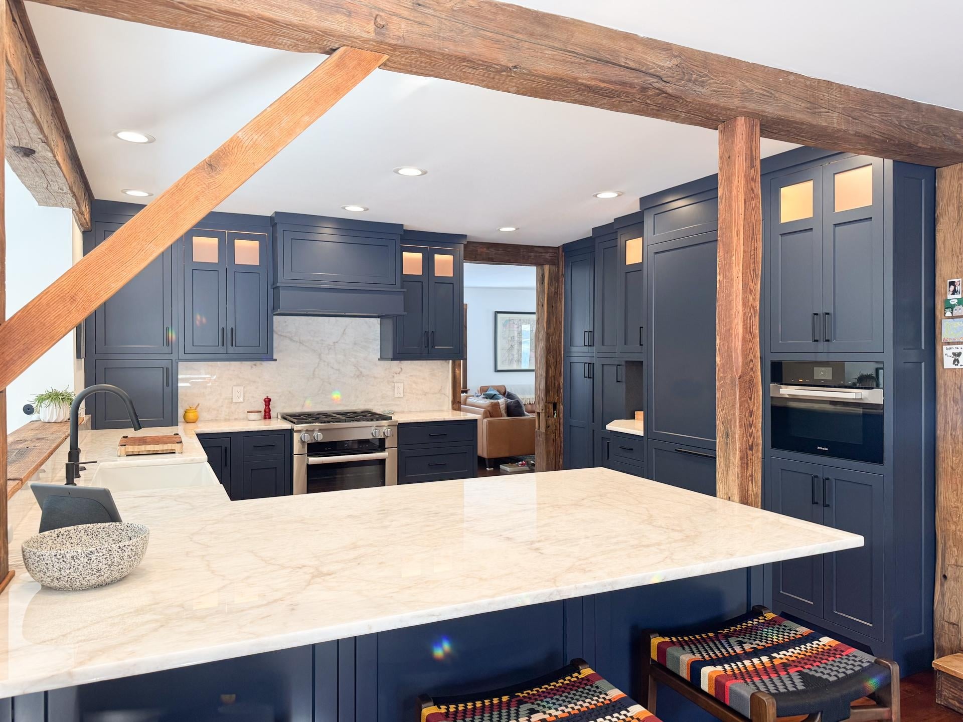 Wide view of the new kitchen with navy cabinets, white marble counters and backsplash, a black faucet, a gas range, and heavy wood beams and posts framing the room.