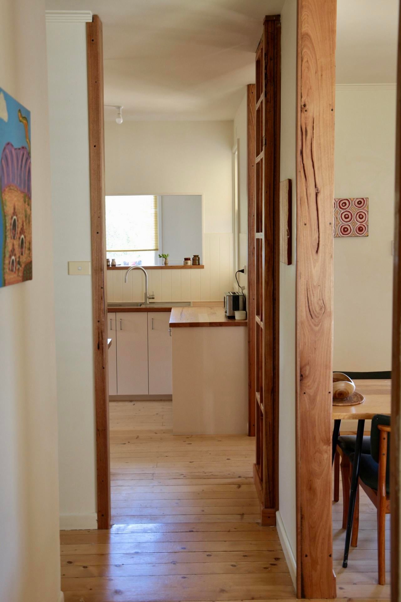 View down a hallway framed by tall timber posts into a bright kitchen with a sink under a window and wood countertops.