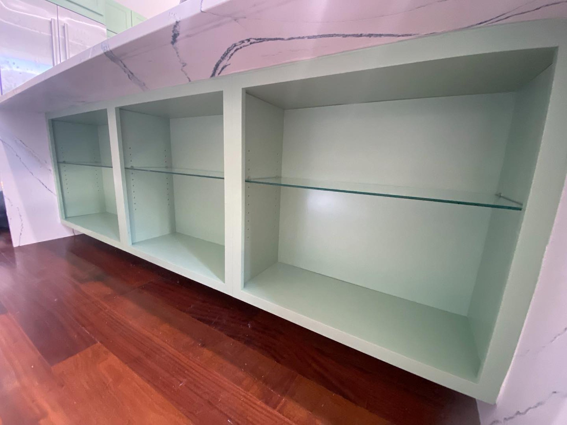 Close-up of the painted island showing three open display shelves in Haven green under a thick Cambria quartz waterfall edge, with cherry wood flooring below.