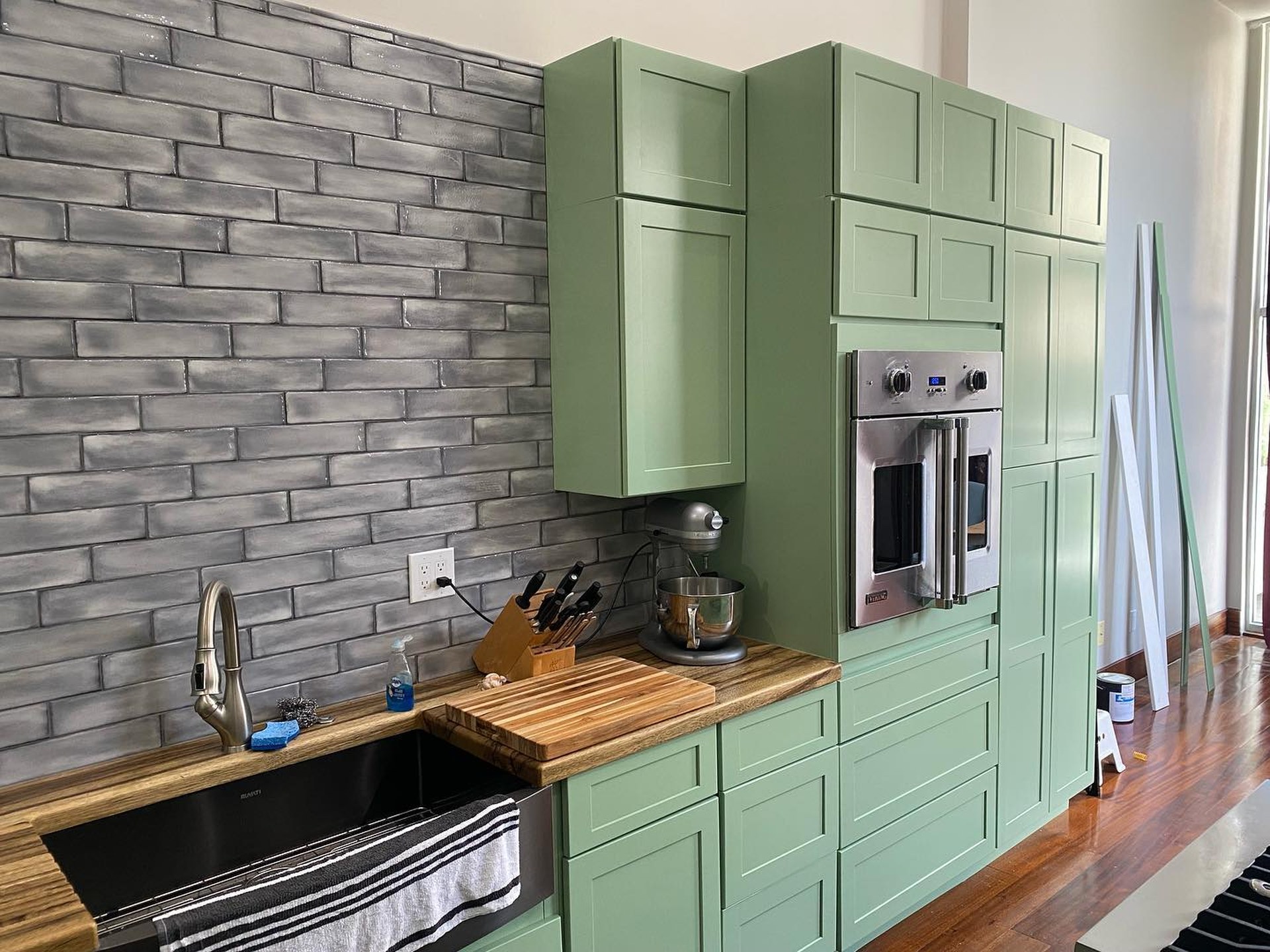 Mid-range view of the painted kitchen showing Haven green cabinets, grey hand-glazed brick backsplash, black apron sink, Viking wall oven, and butcher block counters.