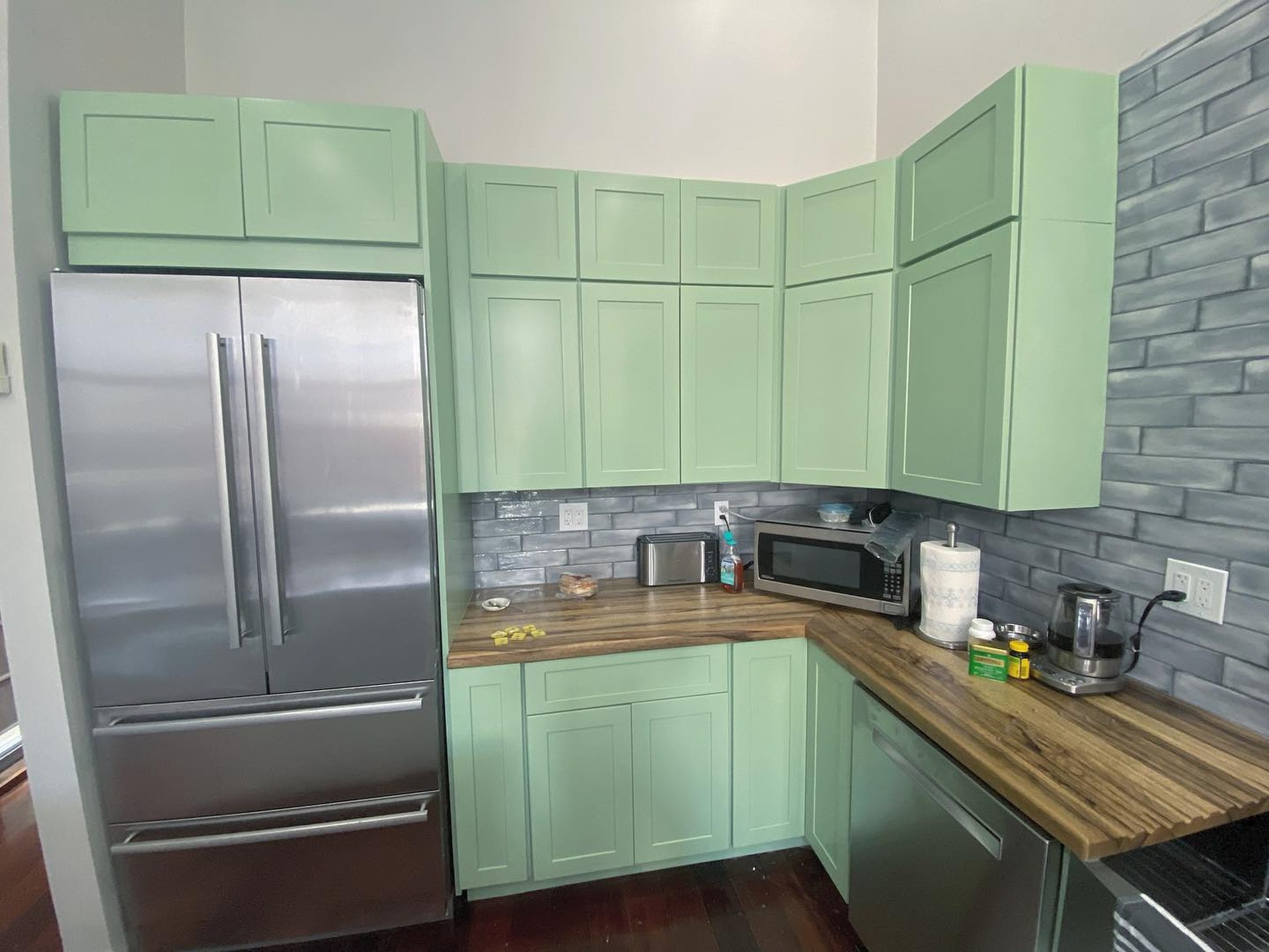 After view of the kitchen fridge corner with Haven green cabinets, a toaster, microwave and kettle on the butcher block counter, grey brick backsplash, and the stainless built-in fridge.