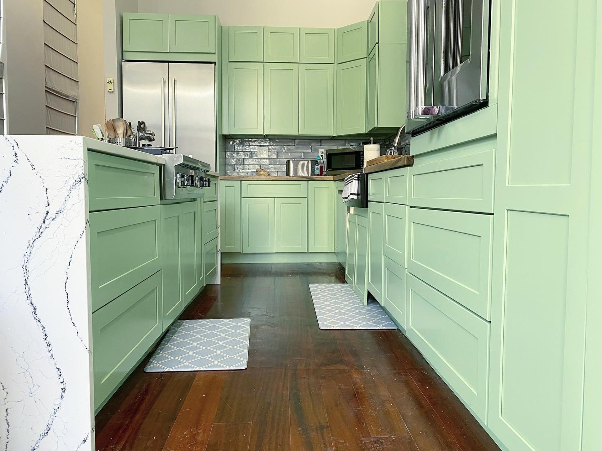 Low angle view of the galley kitchen after painting, with Haven green shaker drawers on both sides, waterfall quartz island at left, and cherry wood floors leading to a corner cabinet run.