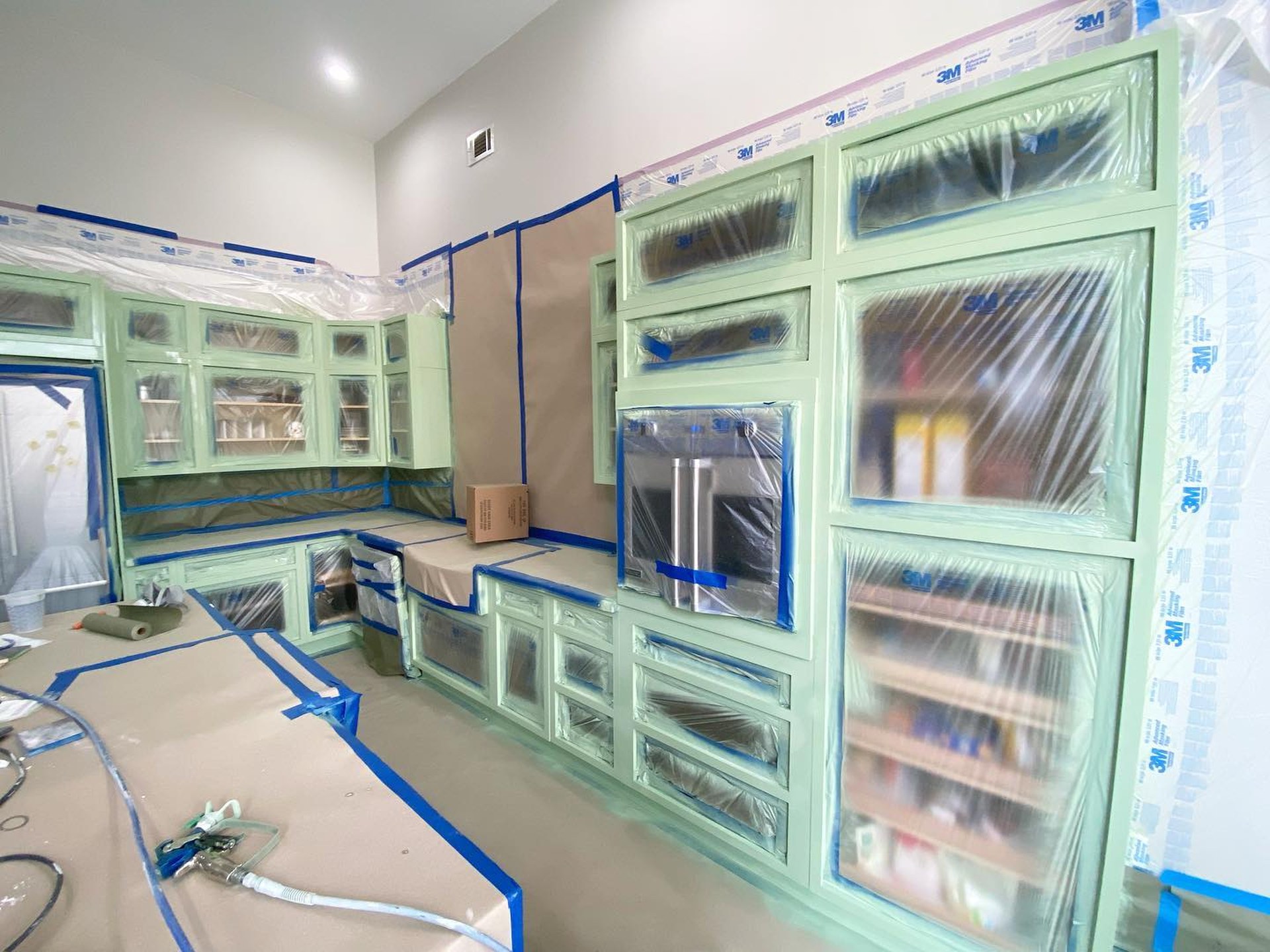Kitchen cabinets masked with blue tape and 3M plastic sheeting mid-project, with the first coats of Sherwin-Williams Haven green visible on cabinet frames and drawer fronts.
