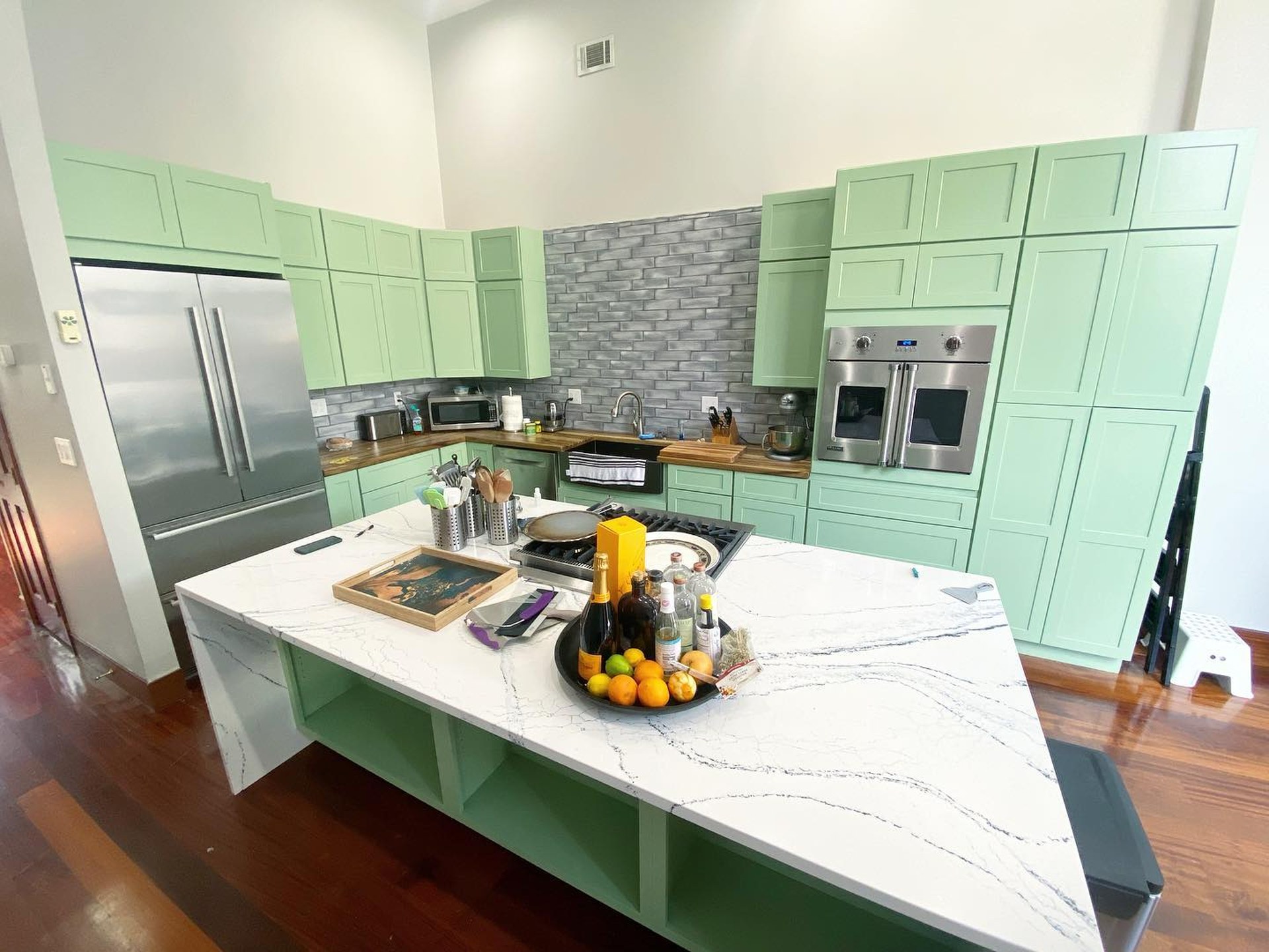 Same wide kitchen view, now with every cabinet painted Sherwin-Williams Haven green against the grey brick backsplash and Cambria quartz waterfall island.