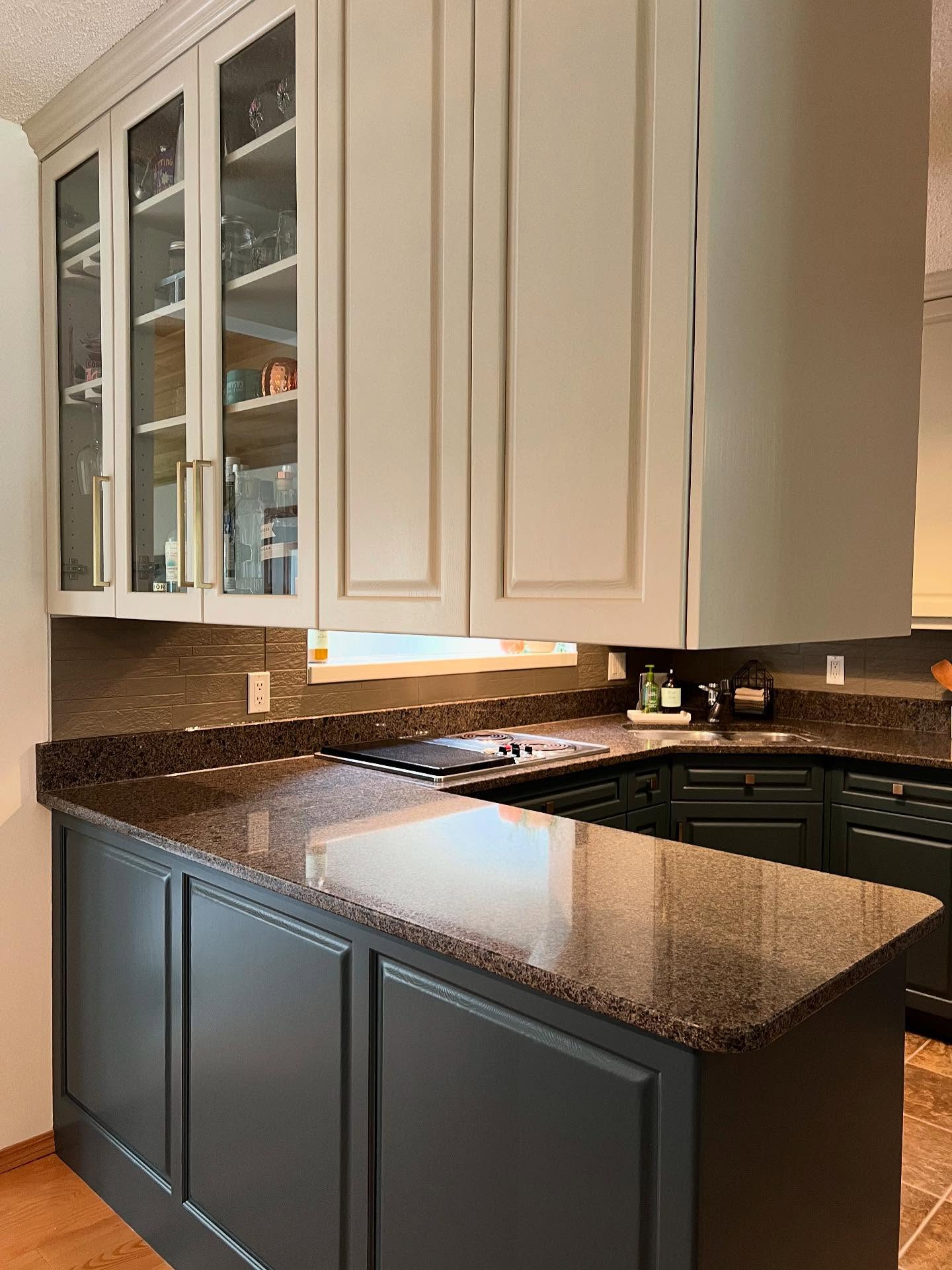 Kitchen peninsula with Hazy Skies sage painted glass-front upper cabinets holding glassware and bottles, Lead Grey dark forest green lower cabinets, long brass bar pulls, speckled granite peninsula counter, a cooktop on the back counter, and a dark painted stone-look backsplash.