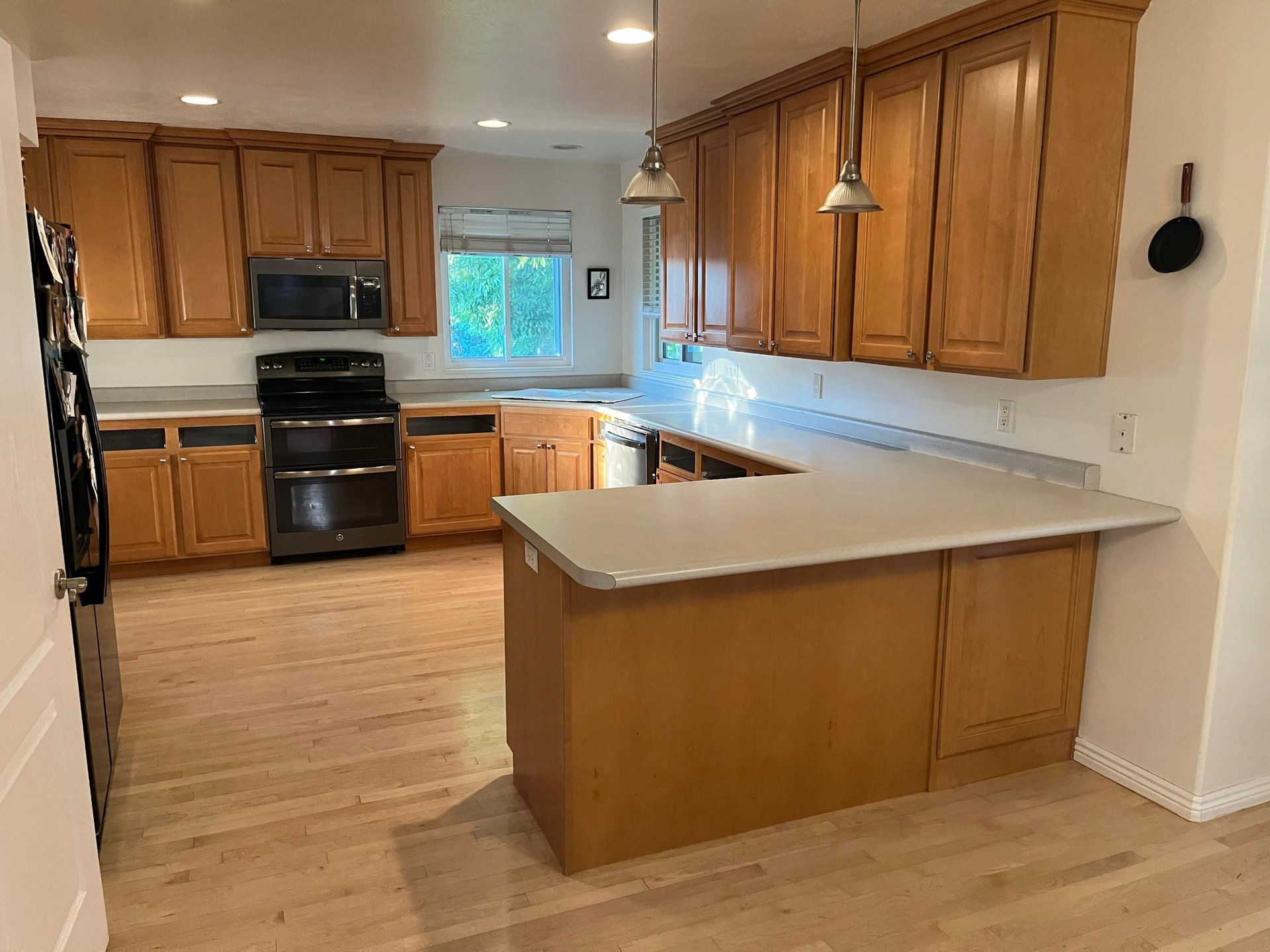 U-shaped kitchen with worn honey oak cabinets, light gray formica countertops, a chipped overcounter sink, black appliances, two silver pendant lights, and bare light wood flooring.