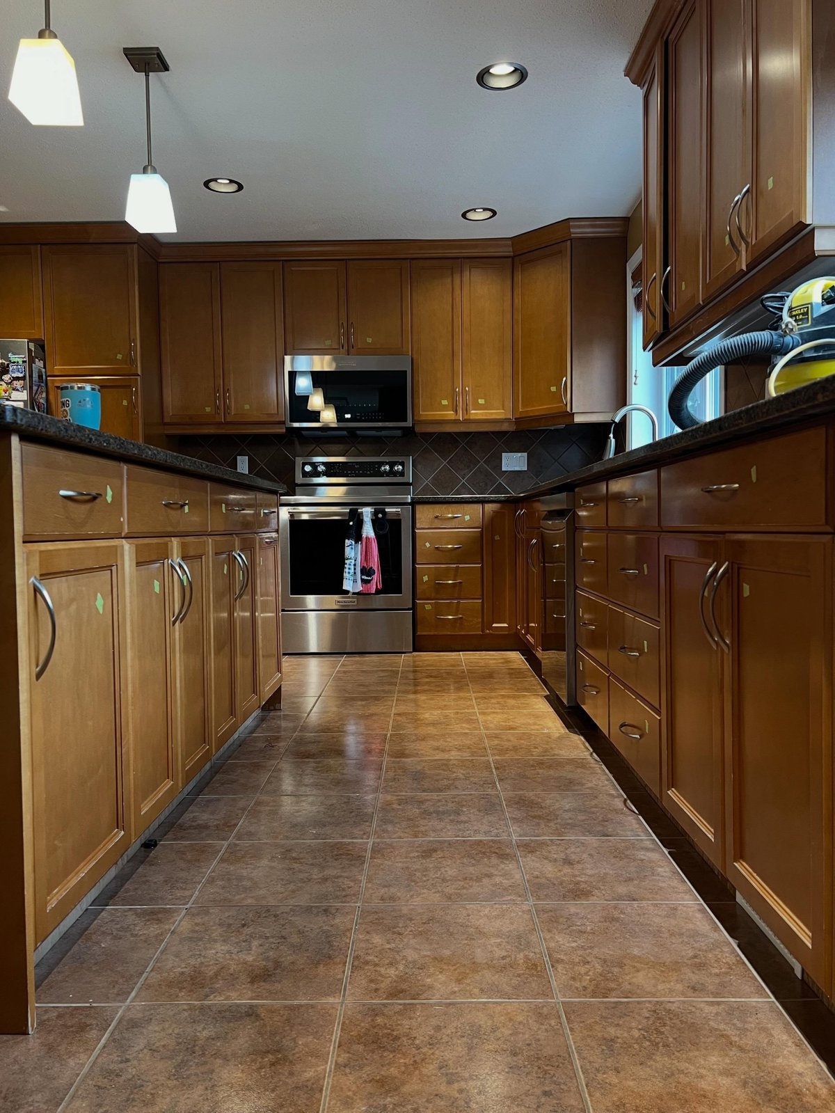 Narrow view down the kitchen galley toward the stainless steel oven, lined with orange honey oak cabinets on both sides and a brown diamond backsplash behind the stove.