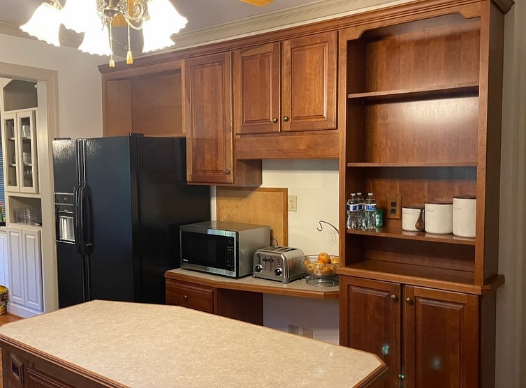 Dated kitchen corner with a black side-by-side fridge, honey oak upper cabinets, a honey oak bookshelf column with three open shelves holding water bottles and canisters, a black Panasonic microwave in a counter nook, and a stainless toaster.