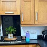 Honey oak shaker kitchen before painting, with a stainless gas hob set into a black laminate counter, a potted plant on a wooden tray, black floor tile and a run of orange-toned oak cabinets filling the frame.