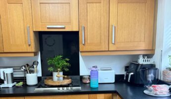 Honey oak shaker kitchen before painting, with a stainless gas hob set into a black laminate counter, a potted plant on a wooden tray, black floor tile and a run of orange-toned oak cabinets filling the frame.