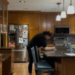 Wide view of a honey oak kitchen before painting, with orange-stained raised-panel cabinets, dark speckled brown granite counters, brown diamond-pattern tile backsplash, warm terracotta tile floor and stainless steel appliances.