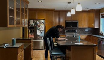 Wide view of a honey oak kitchen before painting, with orange-stained raised-panel cabinets, dark speckled brown granite counters, brown diamond-pattern tile backsplash, warm terracotta tile floor and stainless steel appliances.