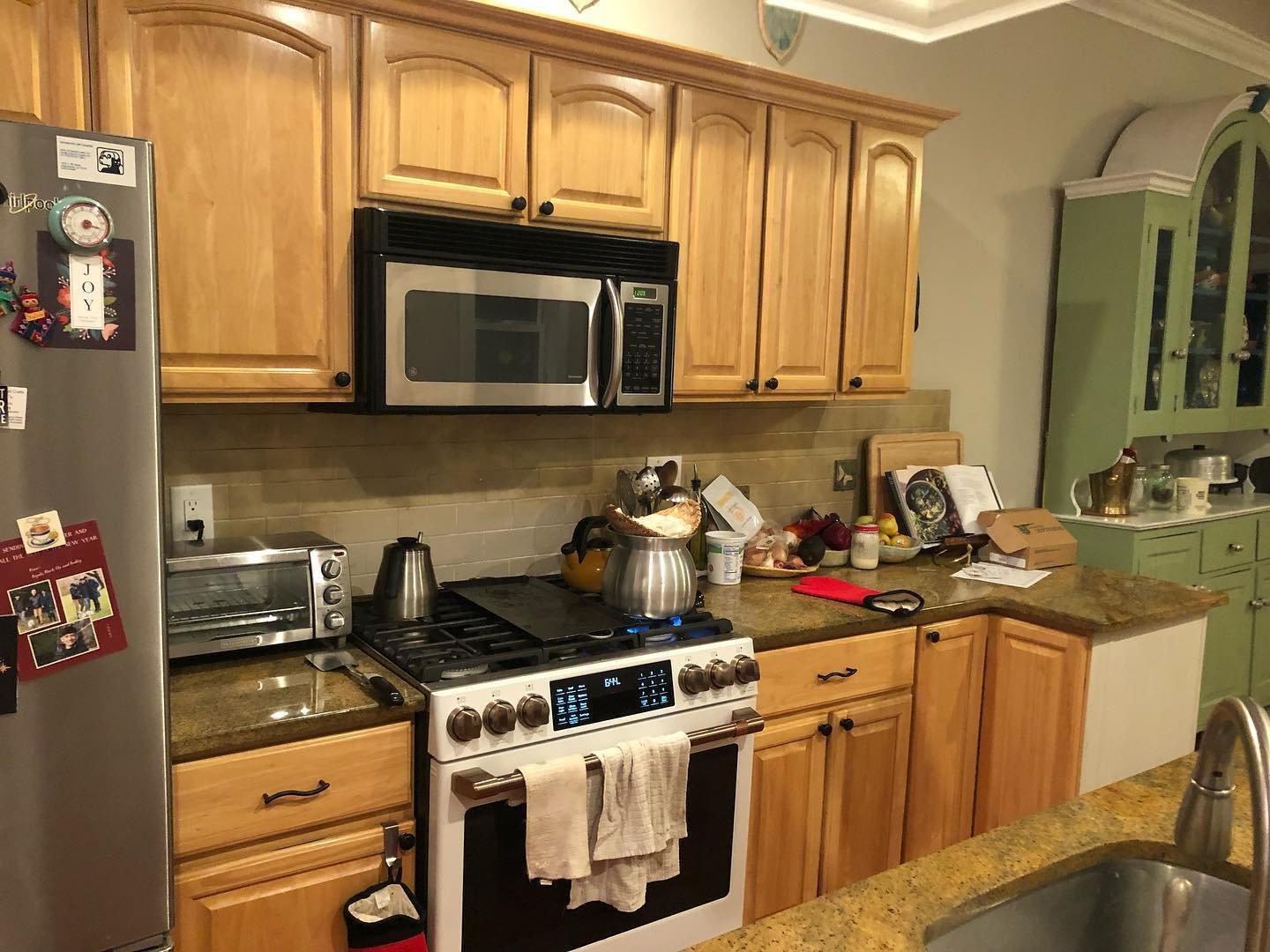 Close-up of a kitchen before renovation with honey oak upper cabinets, cathedral arch doors, black knobs, a stainless microwave over a white range, and gold granite counters.