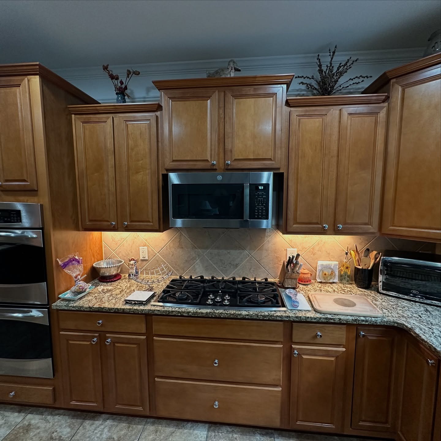 Gas cooktop wall with orange oak upper and lower cabinets, stainless microwave over the stove, double wall ovens on the left, same diamond-set tile backsplash in the background.